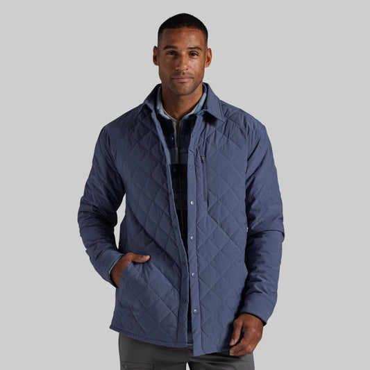 A man wearing a navy blue diamond-quilted button-up jacket over a dark plaid shirt, photographed against a solid light gray background.