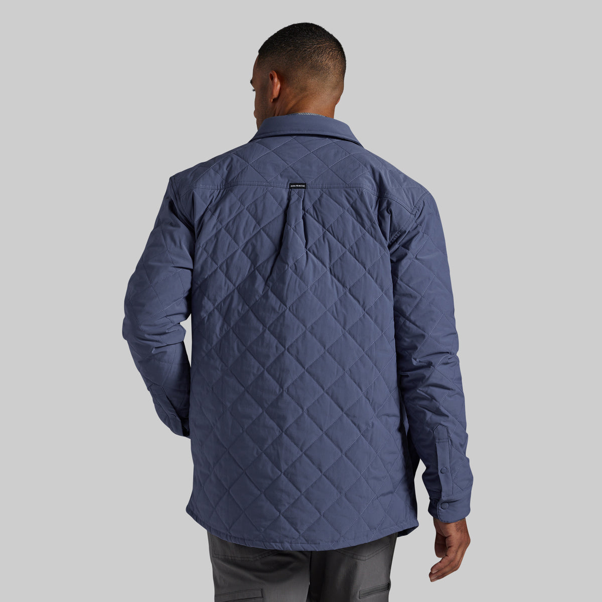 A man viewed from the back wearing a slate blue diamond-quilted jacket, shown against a solid gray studio background.