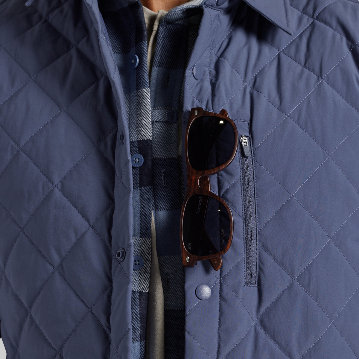 Close-up of a navy blue diamond-quilted jacket with a chest zip pocket, worn over a plaid shirt. A pair of brown sunglasses rests in the pocket.