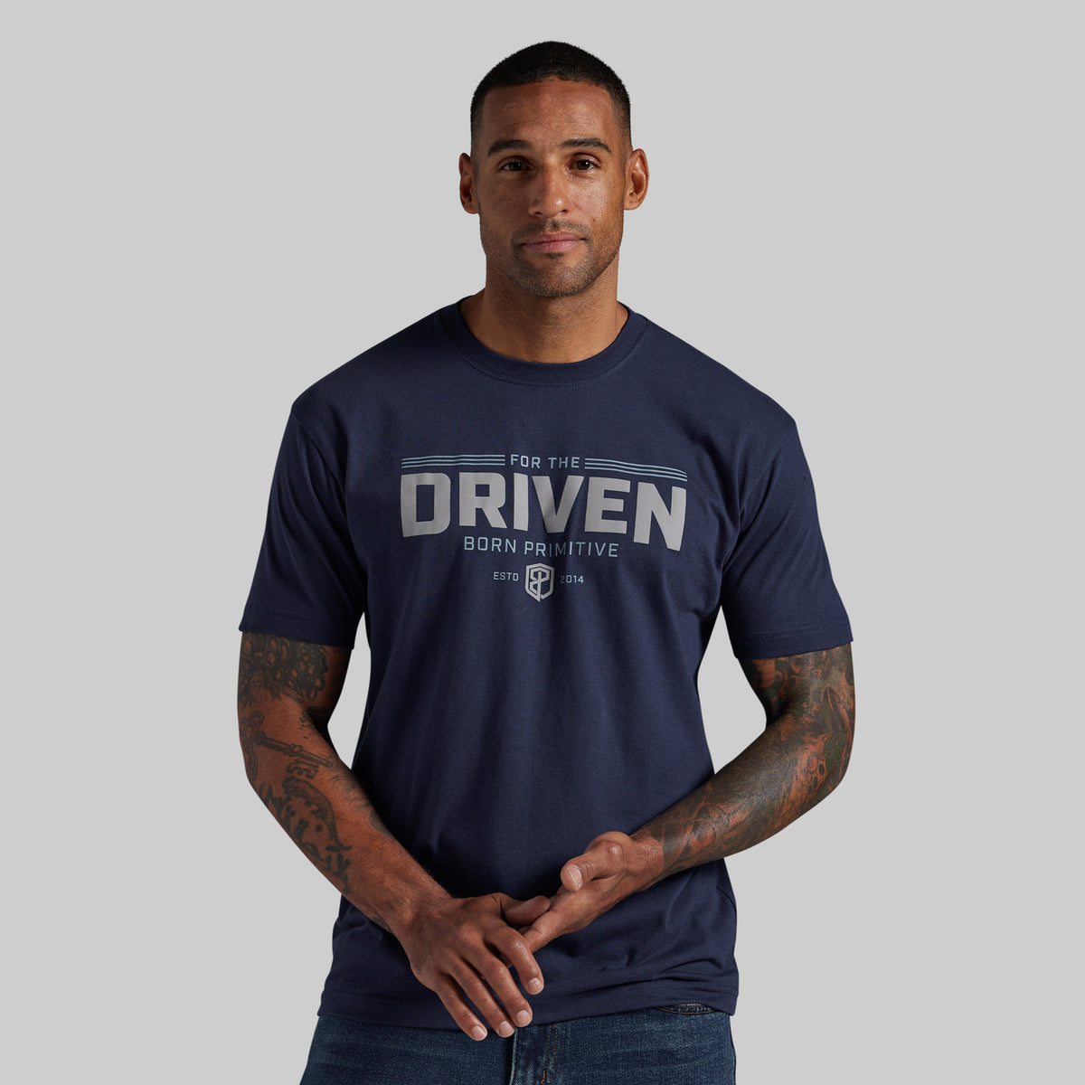A man wearing a navy blue t-shirt with a distressed white graphic that reads 'FOR THE DRIVEN BORN PRIMITIVE' and a shield logo, set against a solid gray background.