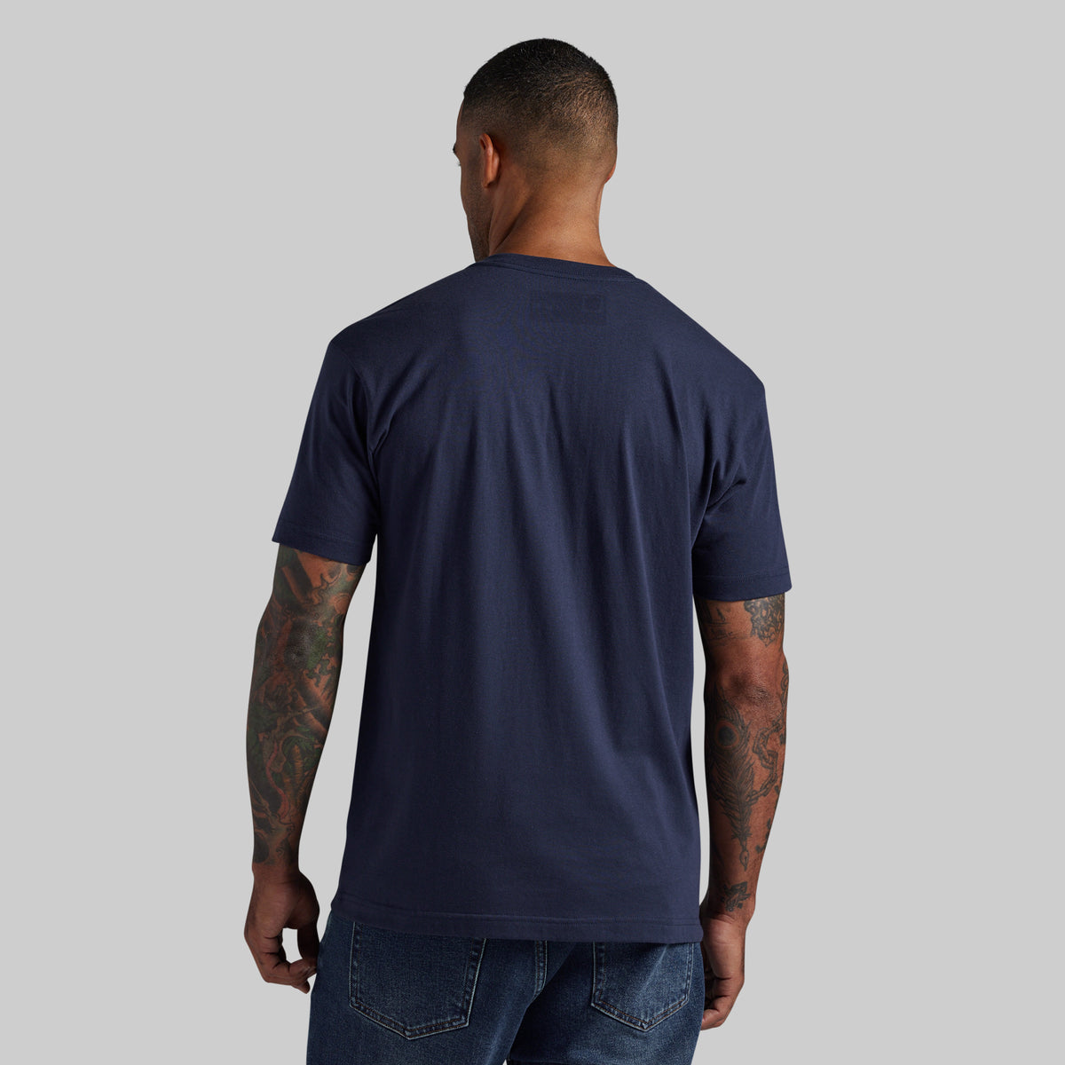 A man viewed from behind wearing a navy blue short-sleeve crewneck t-shirt and blue jeans, standing against a solid light gray background.