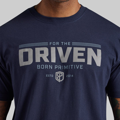 A close-up of a navy blue t-shirt worn by a model, featuring a light gray graphic print on the chest that reads 'FOR THE DRIVEN BORN PRIMITIVE ESTD 2014' with a shield logo.