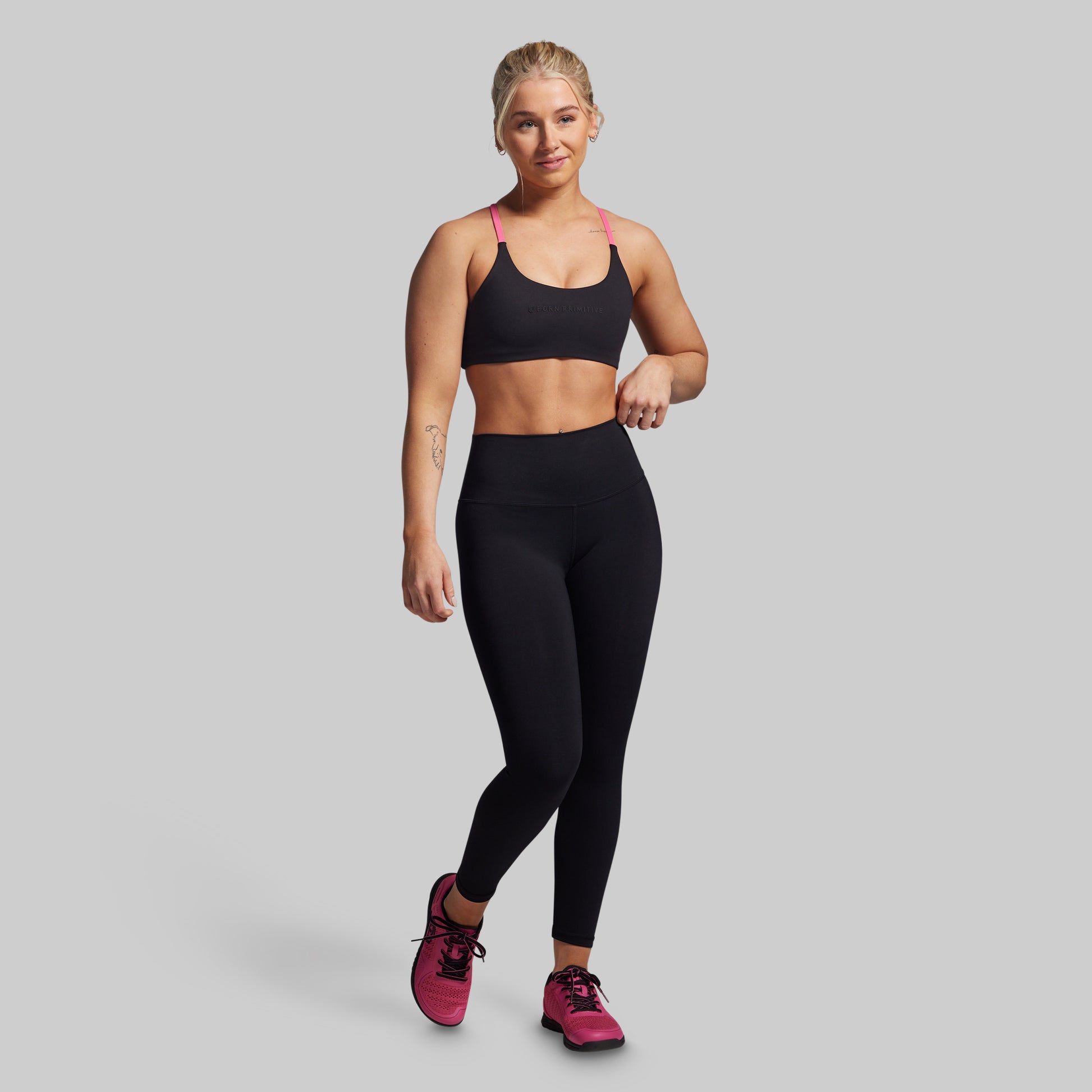 Woman wearing a black sports bra and leggings with pink shoes on a gray background
