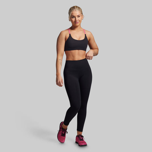 Woman wearing a black sports bra and leggings with pink shoes on a gray background