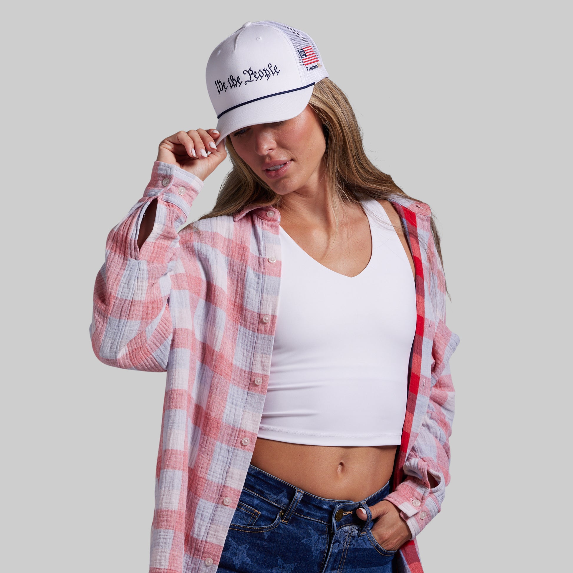 Woman wearing a plaid shirt, white tank top, and jeans with a cap on a plain background