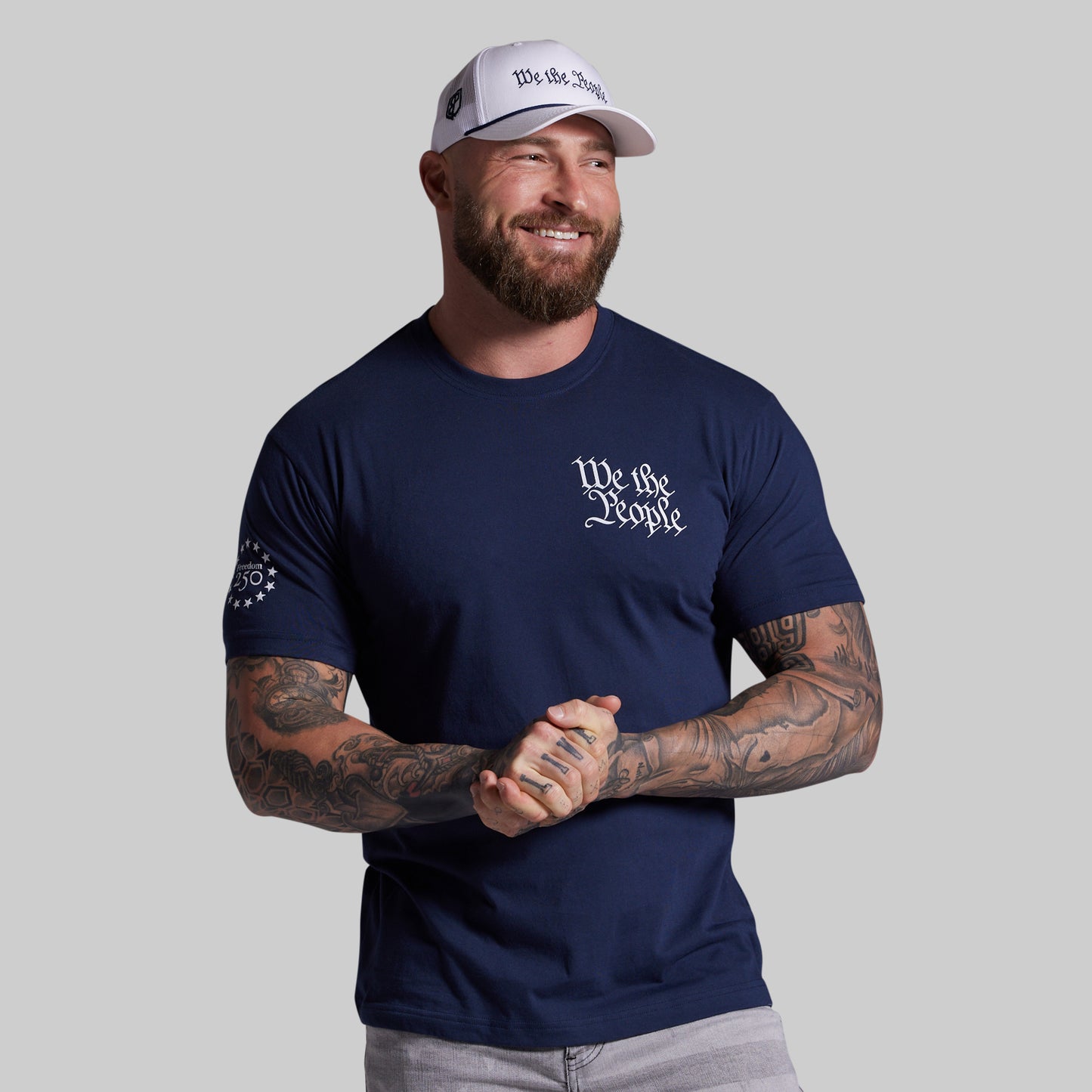 Man wearing a navy blue t-shirt with 'We the People' text and a white cap on a gray background