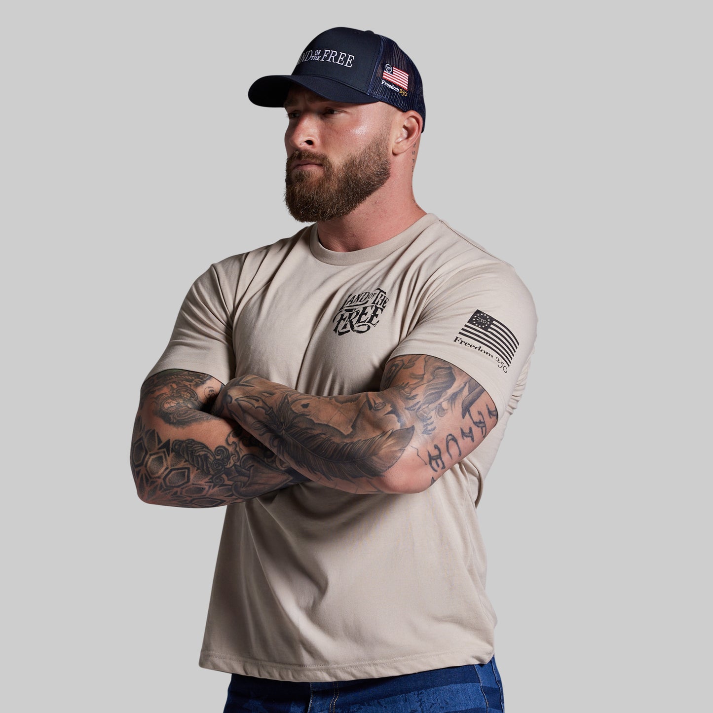 Man wearing a beige t-shirt and navy cap with text and an American flag design, standing against a plain background.