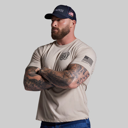 Man wearing a beige t-shirt and navy cap with text and an American flag design, standing against a plain background.
