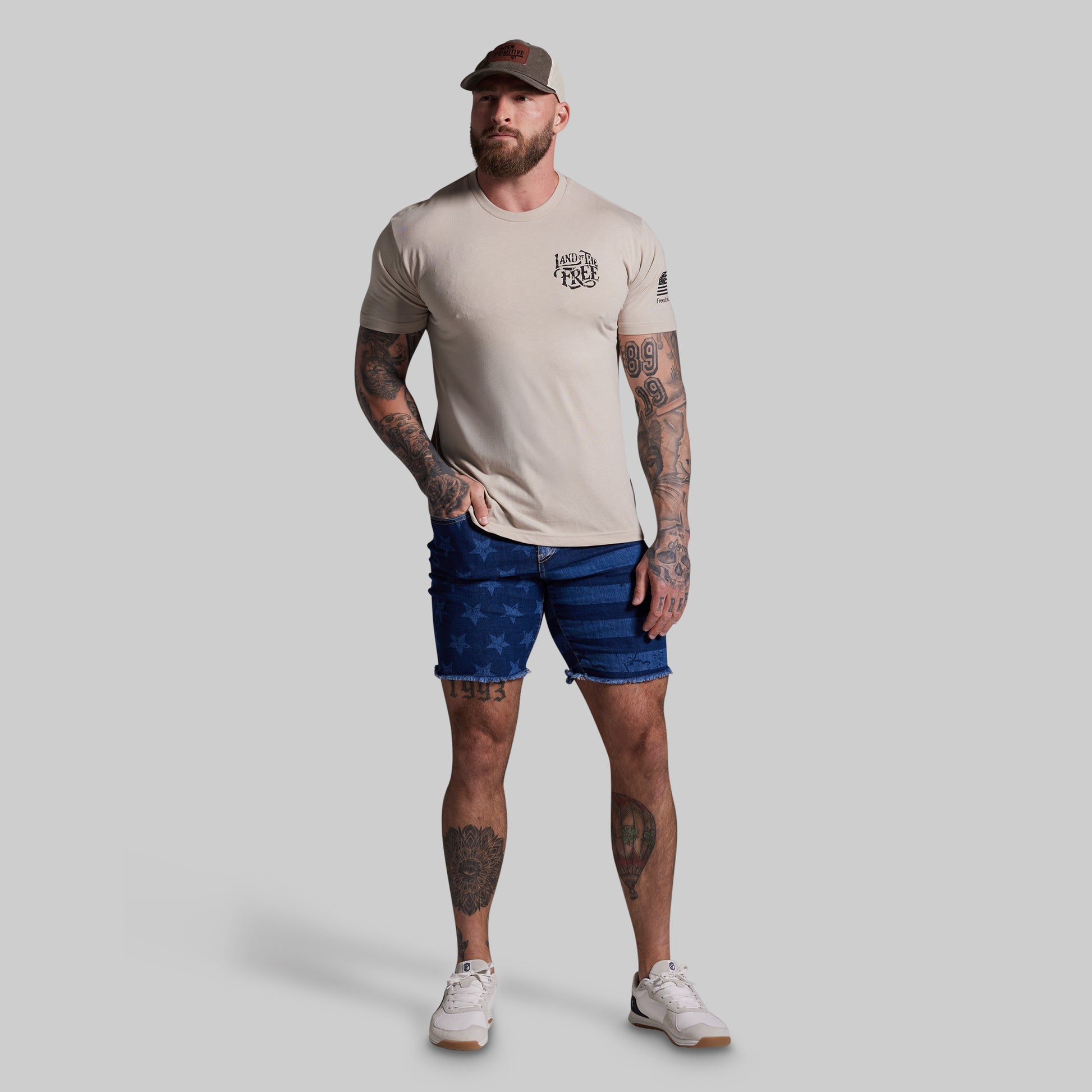 Man wearing a beige t-shirt, blue shorts, and a cap on a gray background