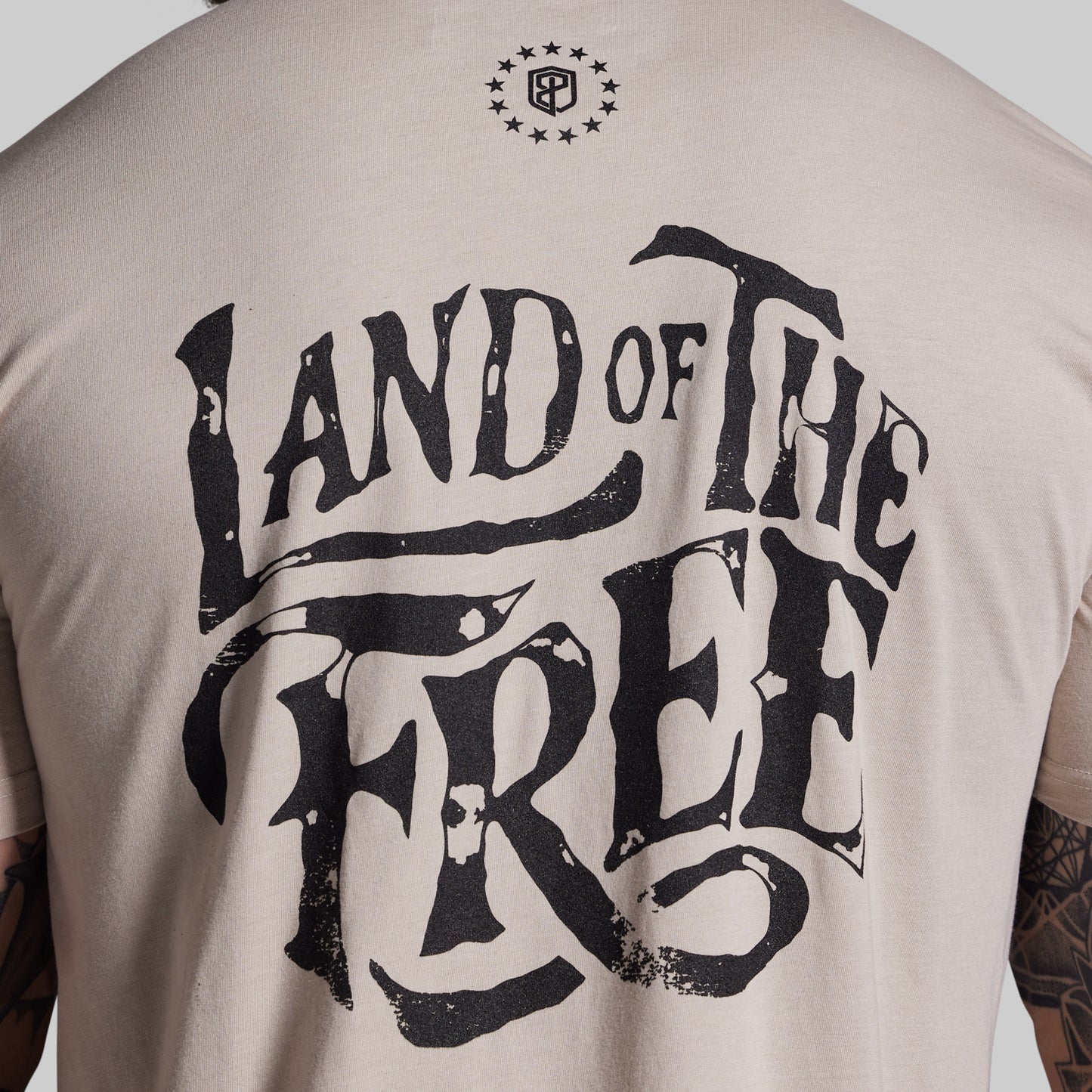 Beige t-shirt with 'Land of the Free' text on the back, worn by a person.