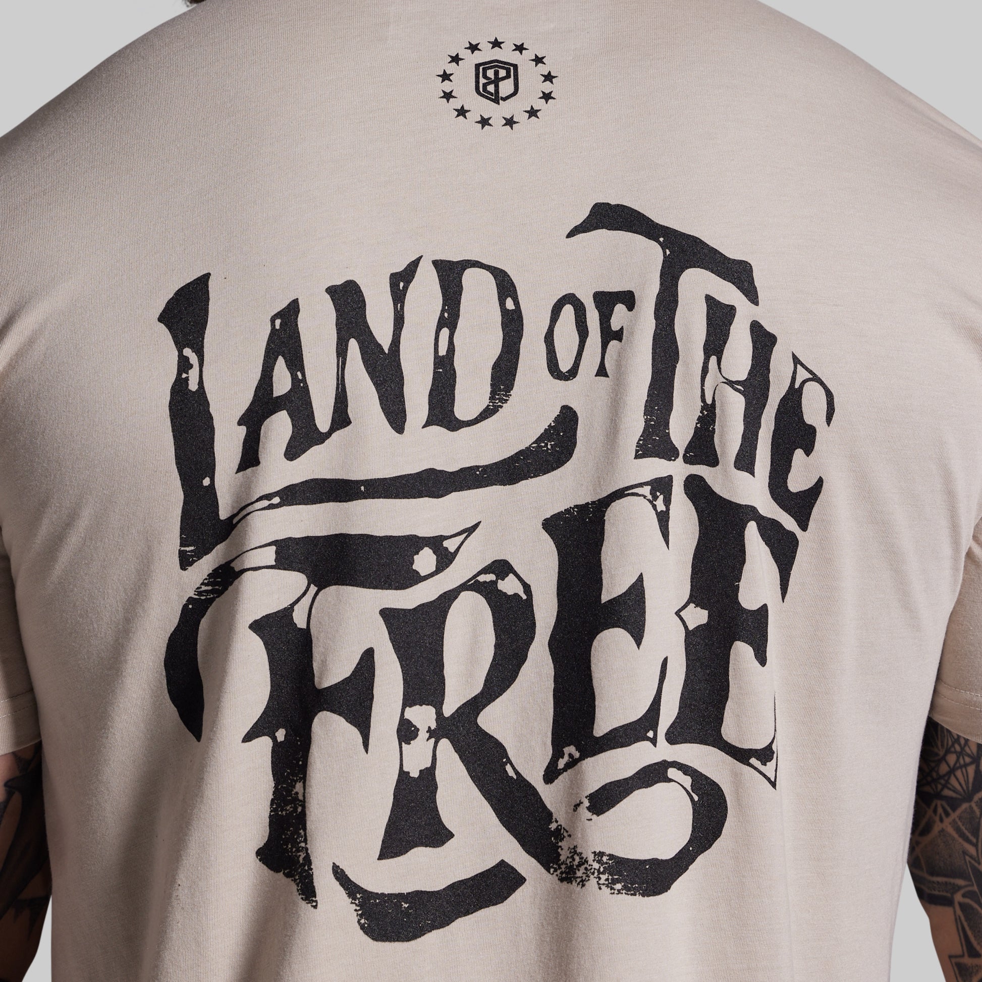 Beige t-shirt with 'Land of the Free' text on the back, worn by a person.