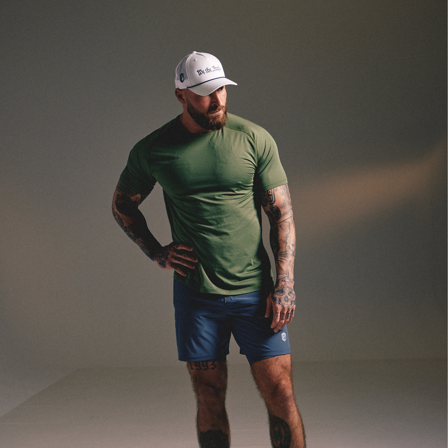 A man modeling a green short-sleeve athletic t-shirt and blue athletic shorts, paired with a white cap, in a studio setting with a warm spotlight on the wall.