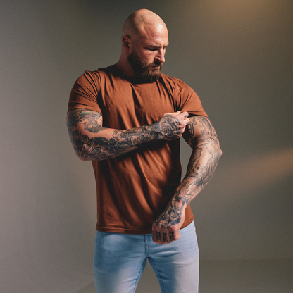A muscular bald man with a beard and extensive tattoos on his arms is wearing a fitted rust-brown t-shirt and light blue jeans, adjusting the sleeve of the shirt against a neutral gray studio background.