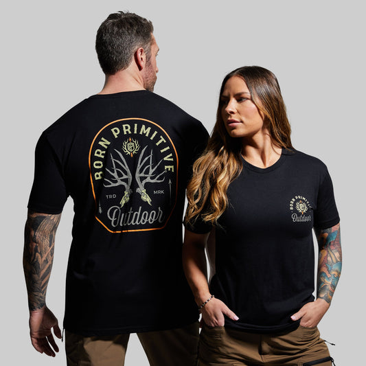 Man and woman wearing black t-shirts with 'Born Primitive' logo on a gray background