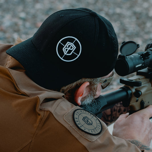 Person wearing a black cap and tan long sleeve with a logo, looking through scope.