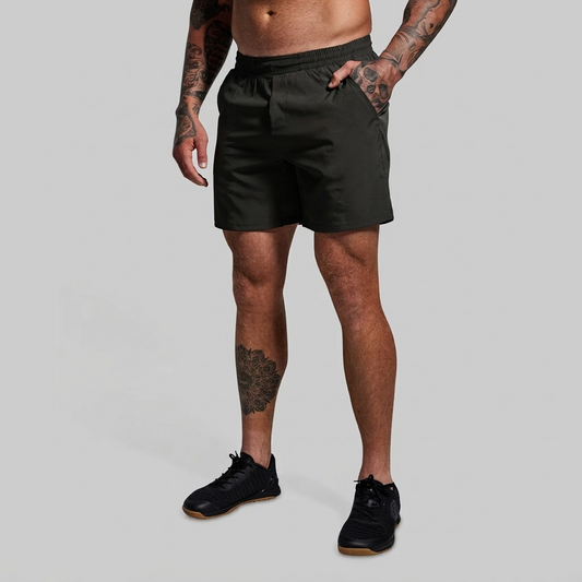 A man wearing dark olive green athletic shorts and black sneakers, shown from the waist down against a solid light gray background.