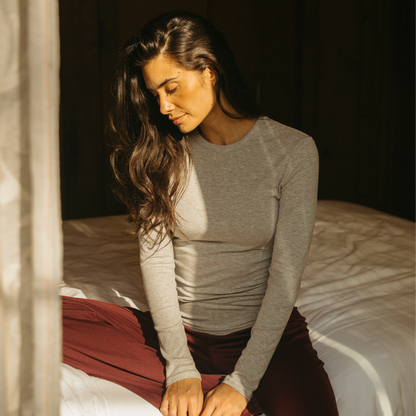 A woman with long dark hair sits on a bed in warm sunlight, wearing a fitted heather gray long-sleeve top and burgundy pants.