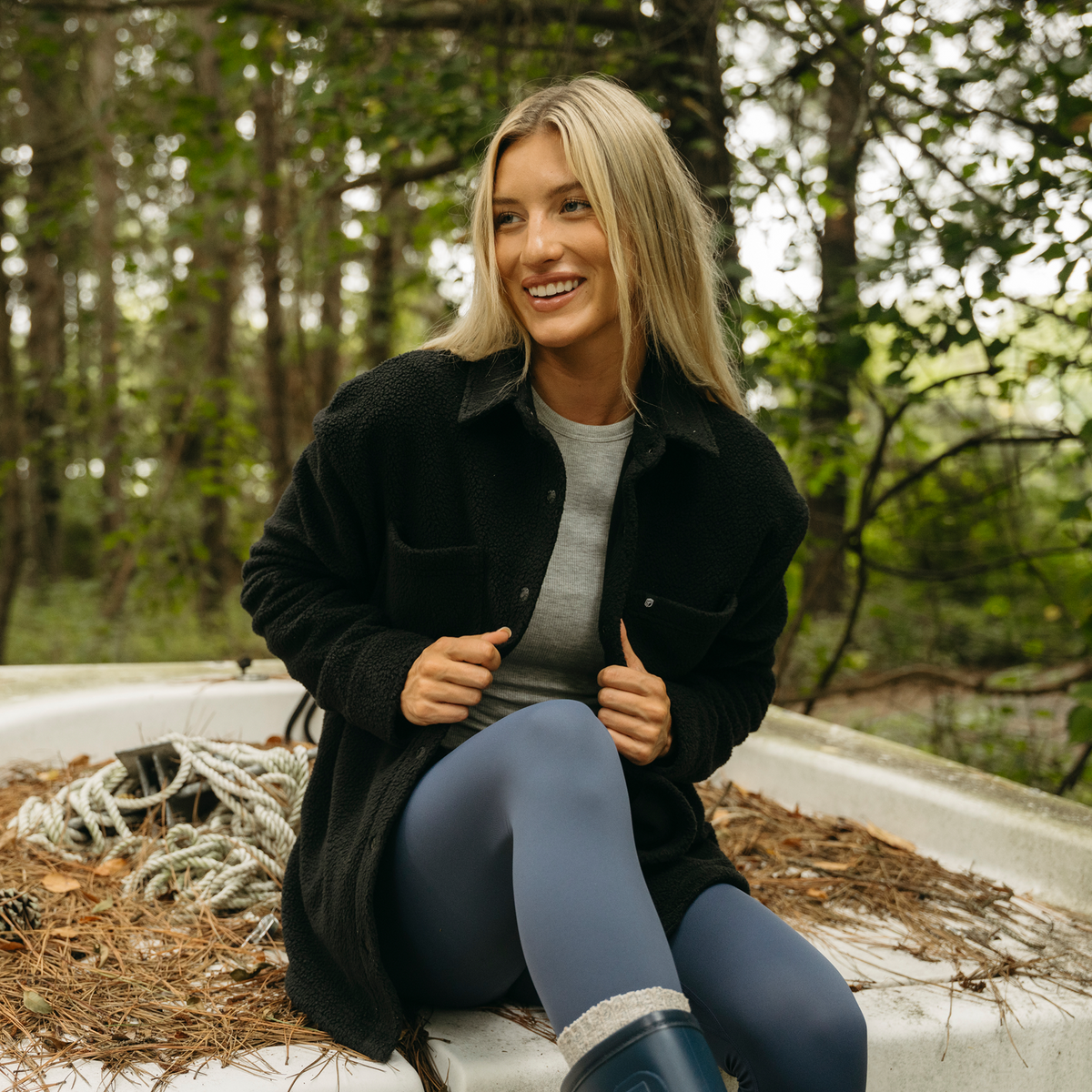 A woman is wearing a black button-up shirt jacket over a gray t-shirt, blue leggings, and a dark rain boot.