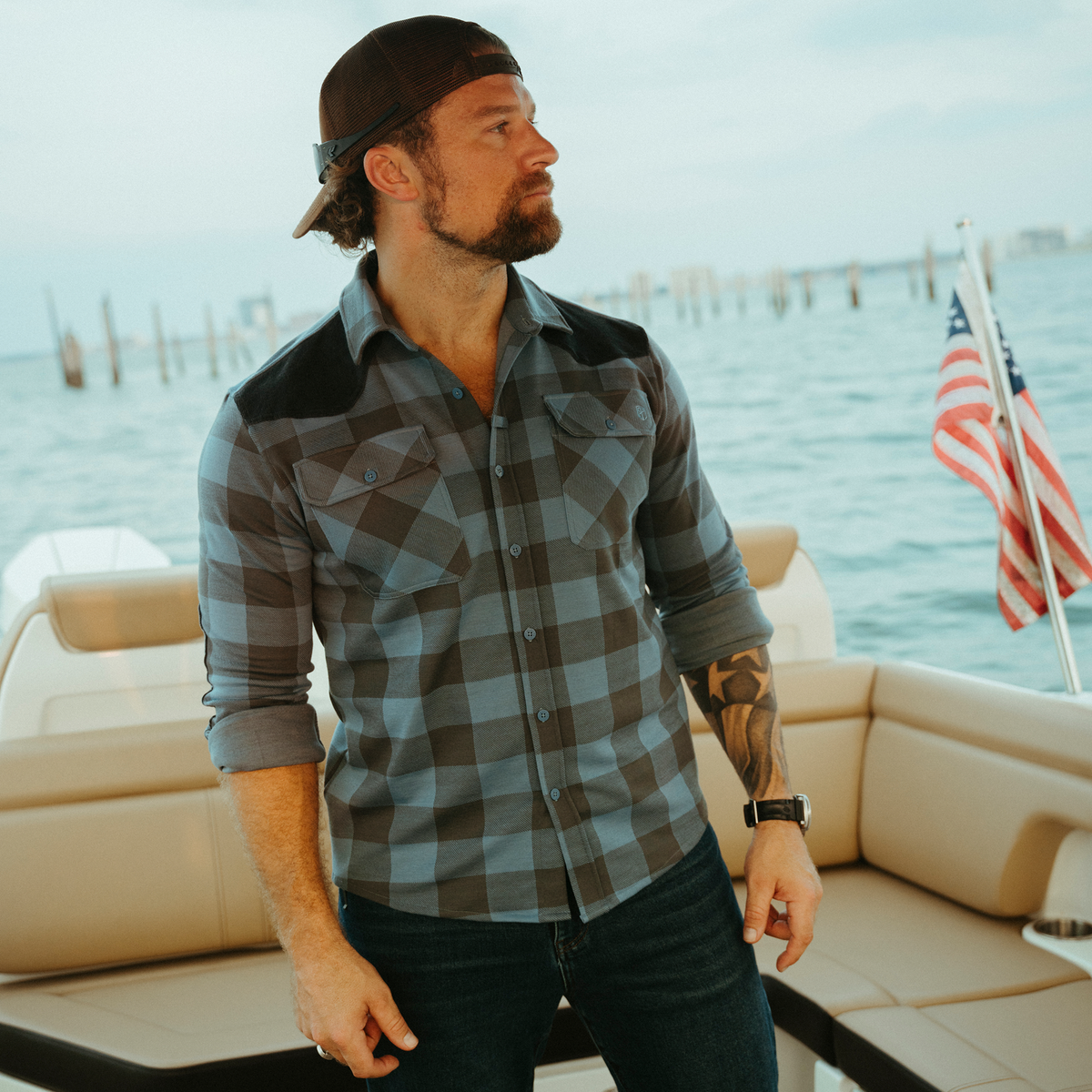 A man wearing a gray and black buffalo plaid button-down shirt with dark shoulder panels, standing on a boat with the ocean and an American flag in the background.
