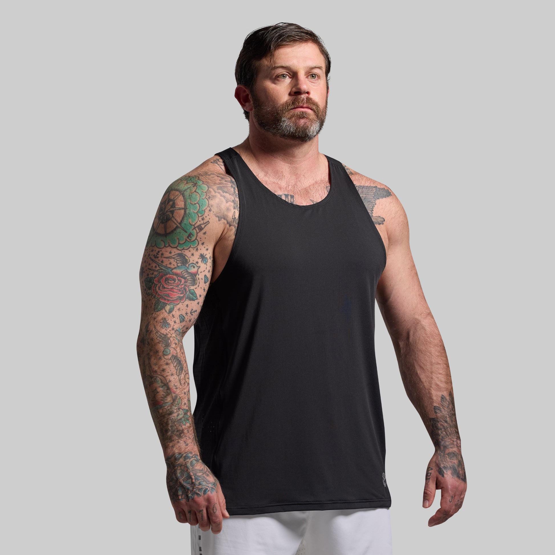 Man wearing a black tank top with tattoos on arms against a gray background