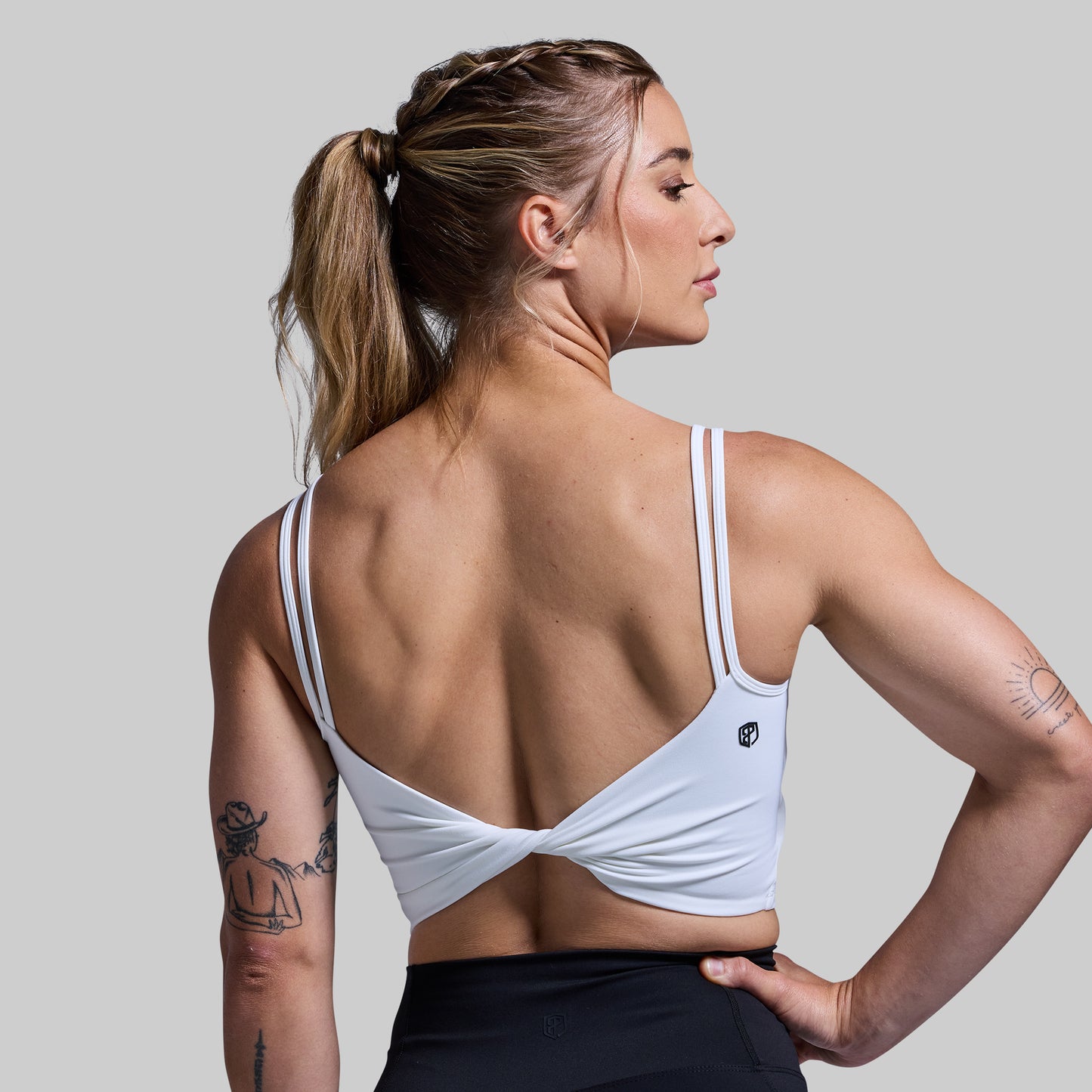 A woman wearing a white twisted sports bra with straps, from the back, with her hand on her hip.