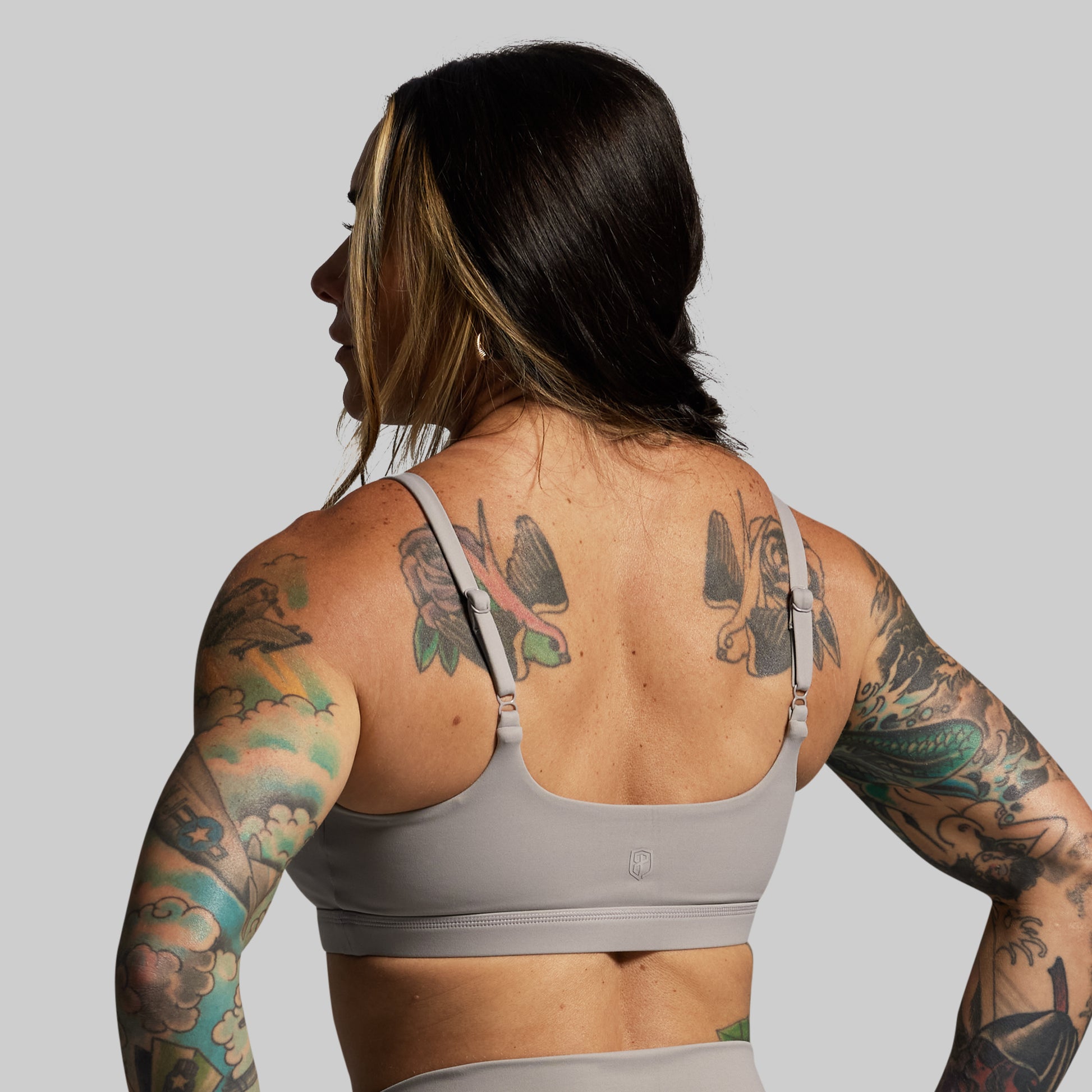 Person wearing a gray sports bra with tattoos on arms and back against a plain background