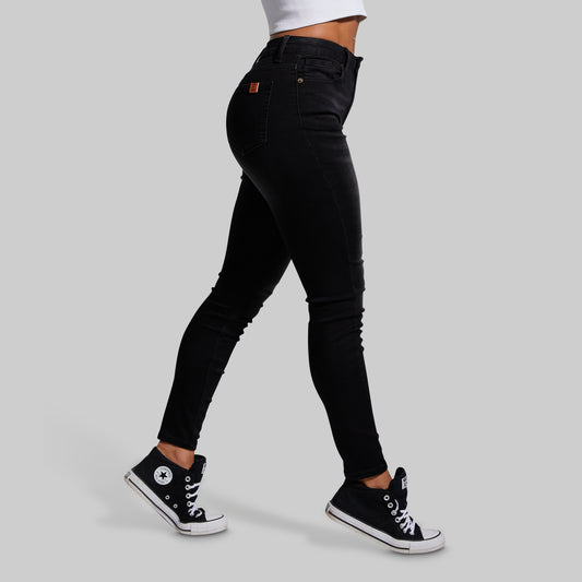 A person wearing black slim-fit jeans and black high-top sneakers, shown from the waist down in a side-profile walking pose against a light gray studio background.