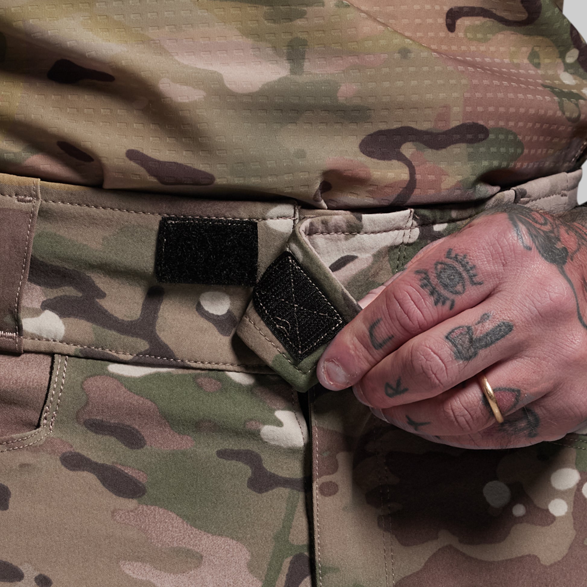 Close-up of camouflage pants with a velcro closure and a hand with tattoos holding the fabric.
