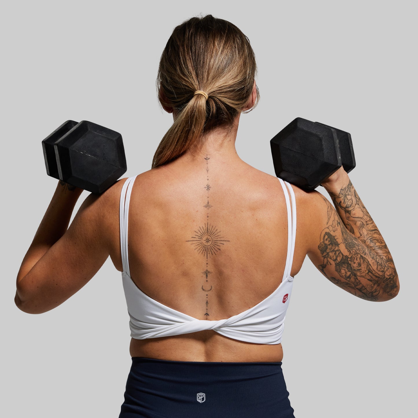 An image of a person wearing a white sports bra with a twisted back design, performing a workout with dumbbells.