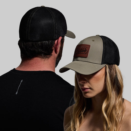 A man wearing a black trucker hat and a woman wearing a green and black trucker hat with a leather patch on it, standing close together facing away from the camera.