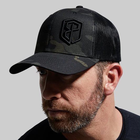 A person wearing a black multicam trucker hat with a camouflage pattern and a logo on the front.
