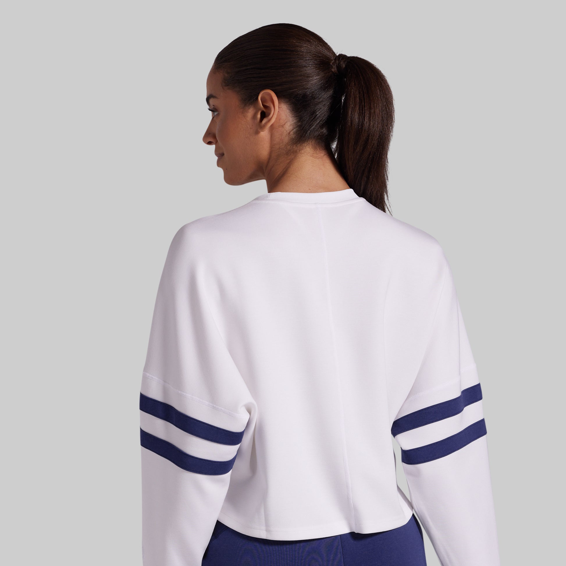 A woman shown from the back wearing a white cropped long-sleeve sweatshirt with navy blue stripes on the sleeves, set against a solid light gray background.