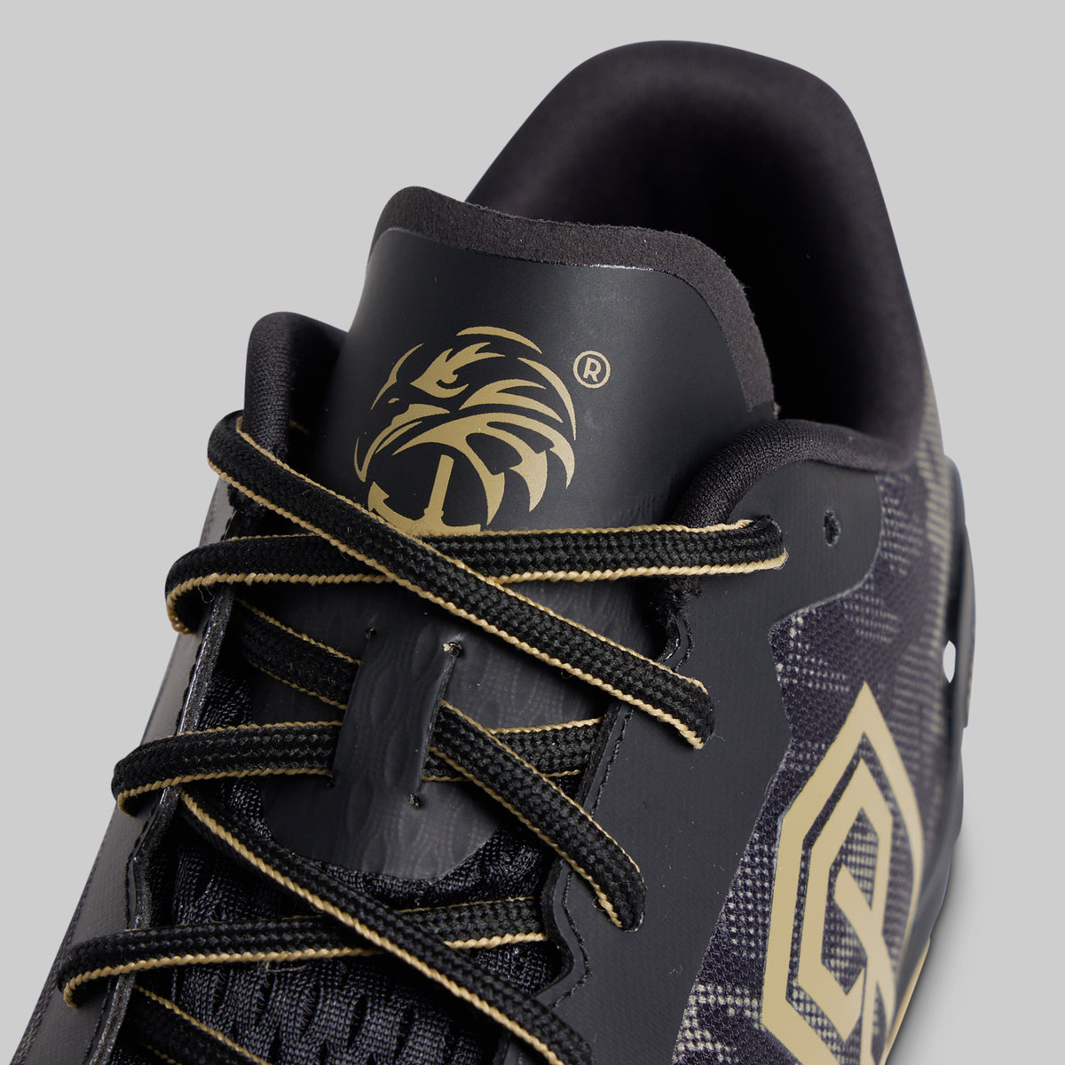 Close-up of a black athletic shoe with gold accents, featuring a gold eagle head logo on the tongue and a large gold geometric emblem on the side, set against a solid gray background.