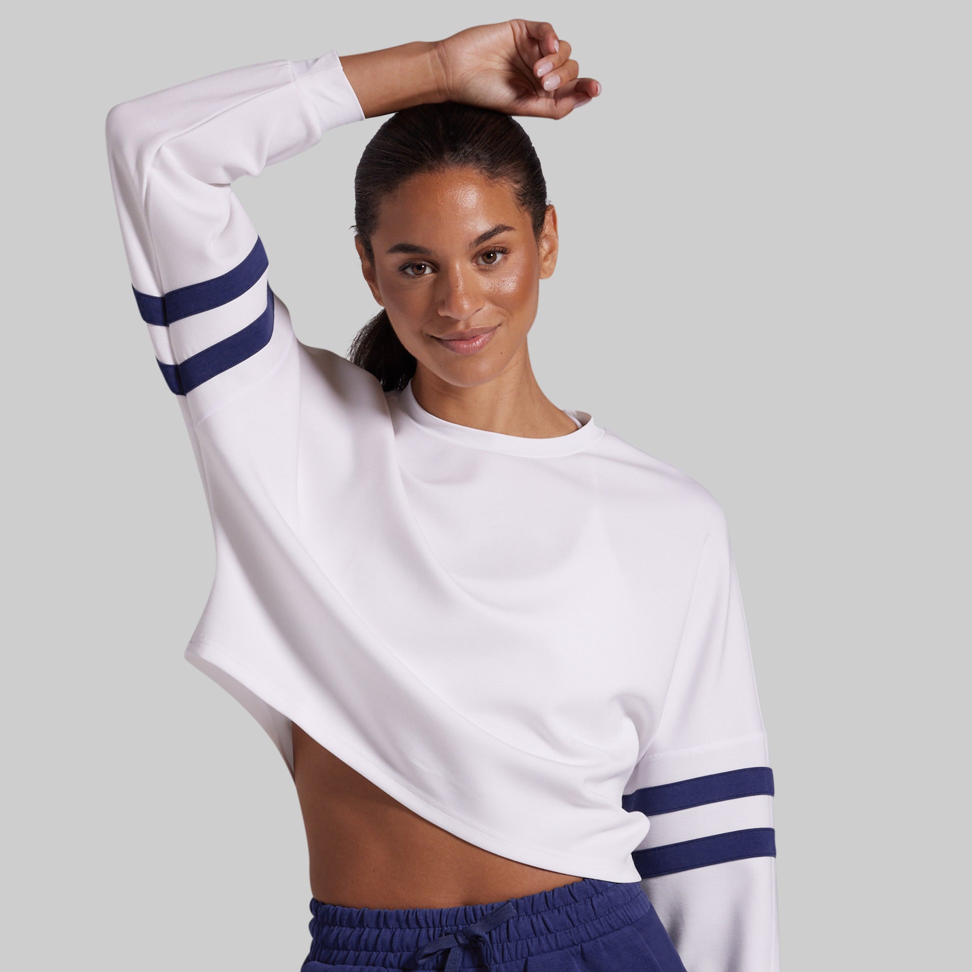 A woman modeling a white cropped long-sleeve sweatshirt with navy blue stripes on the sleeves, paired with navy drawstring lounge shorts, against a solid light gray background.