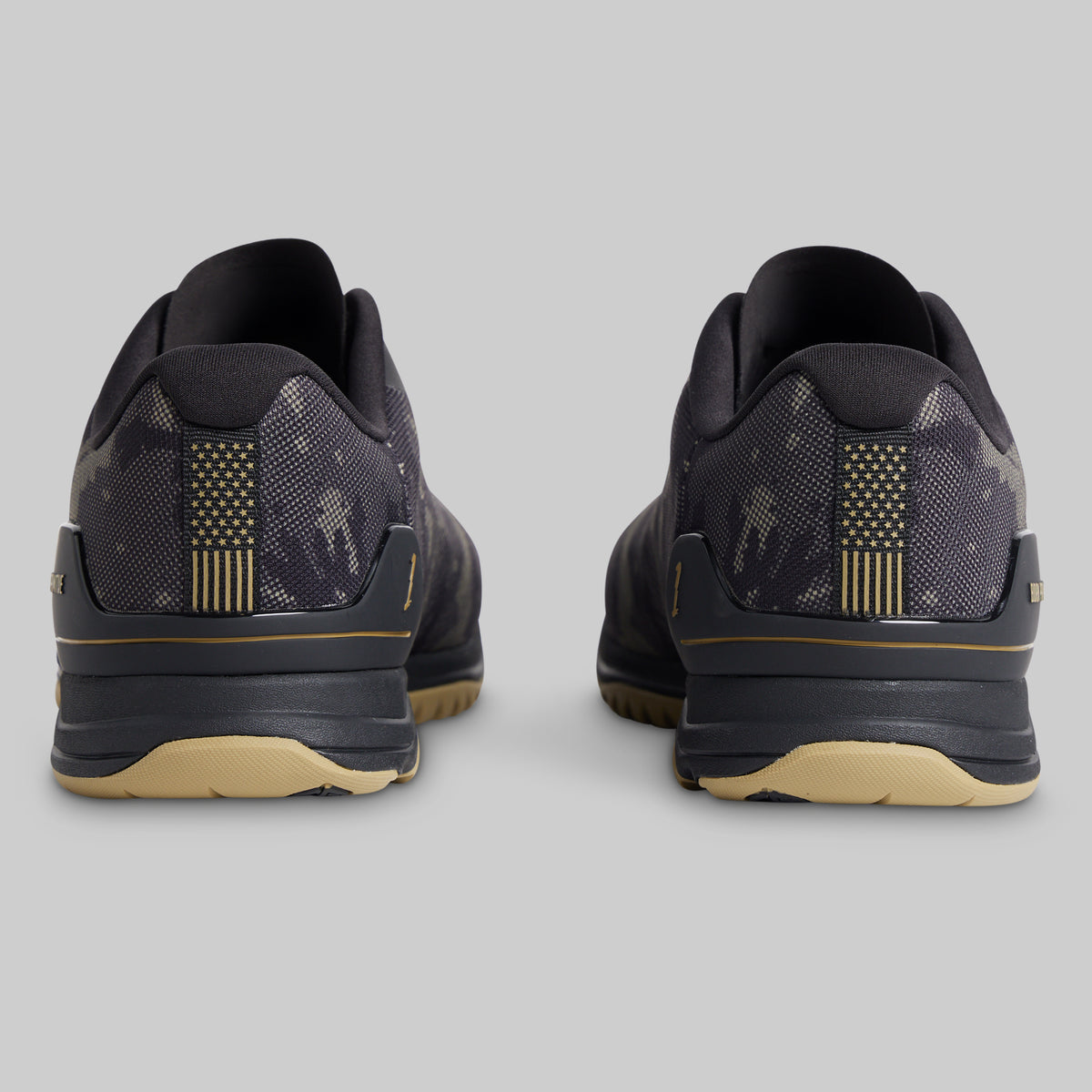A pair of athletic sneakers shown from the rear, featuring a dark gray and black camouflage pattern with gold accents on the heel stripes and midsole, and a beige outsole.