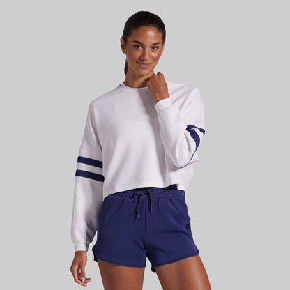 A woman modeling a white long-sleeve cropped sweatshirt with navy blue stripes on the sleeves, paired with navy blue lounge shorts, against a solid gray background.