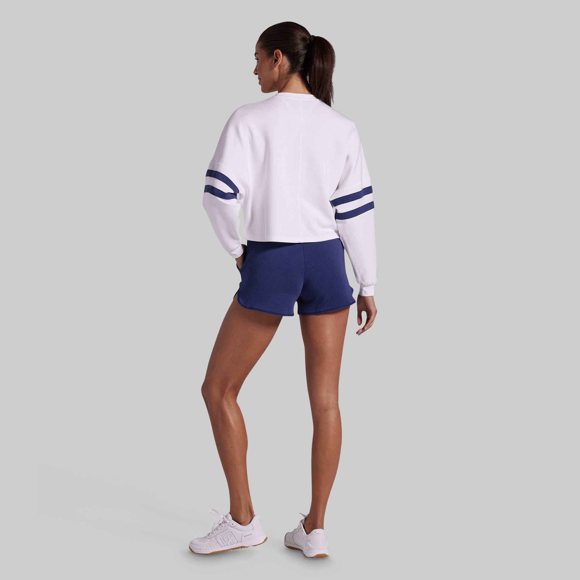 A woman is shown from the back wearing a white long-sleeve cropped sweatshirt with navy blue stripes on the sleeves, navy blue lounge shorts, and white sneakers, set against a solid light gray background.