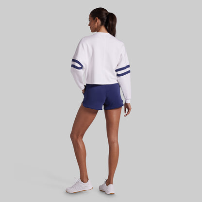 A woman is shown from the back wearing a white long-sleeve cropped sweatshirt with navy blue stripes on the sleeves, navy blue lounge shorts, and white sneakers, set against a solid light gray background.