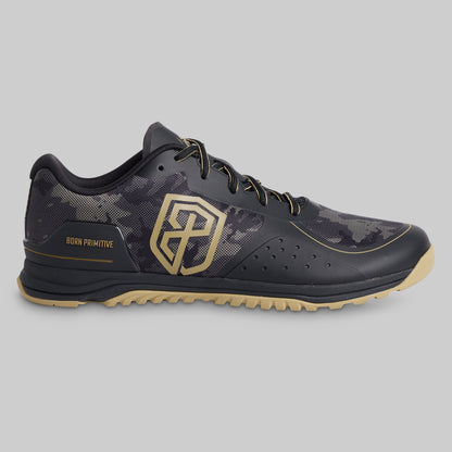 A single Primitives athletic shoe in black and dark gray camouflage with gold accents, including a large geometric logo on the side and 'BORN PRIMITIVE' text, shown against a solid gray background.