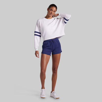 A woman modeling a white cropped long-sleeve sweatshirt with navy blue stripes on the sleeves, paired with navy blue lounge shorts and white sneakers, against a solid gray background.