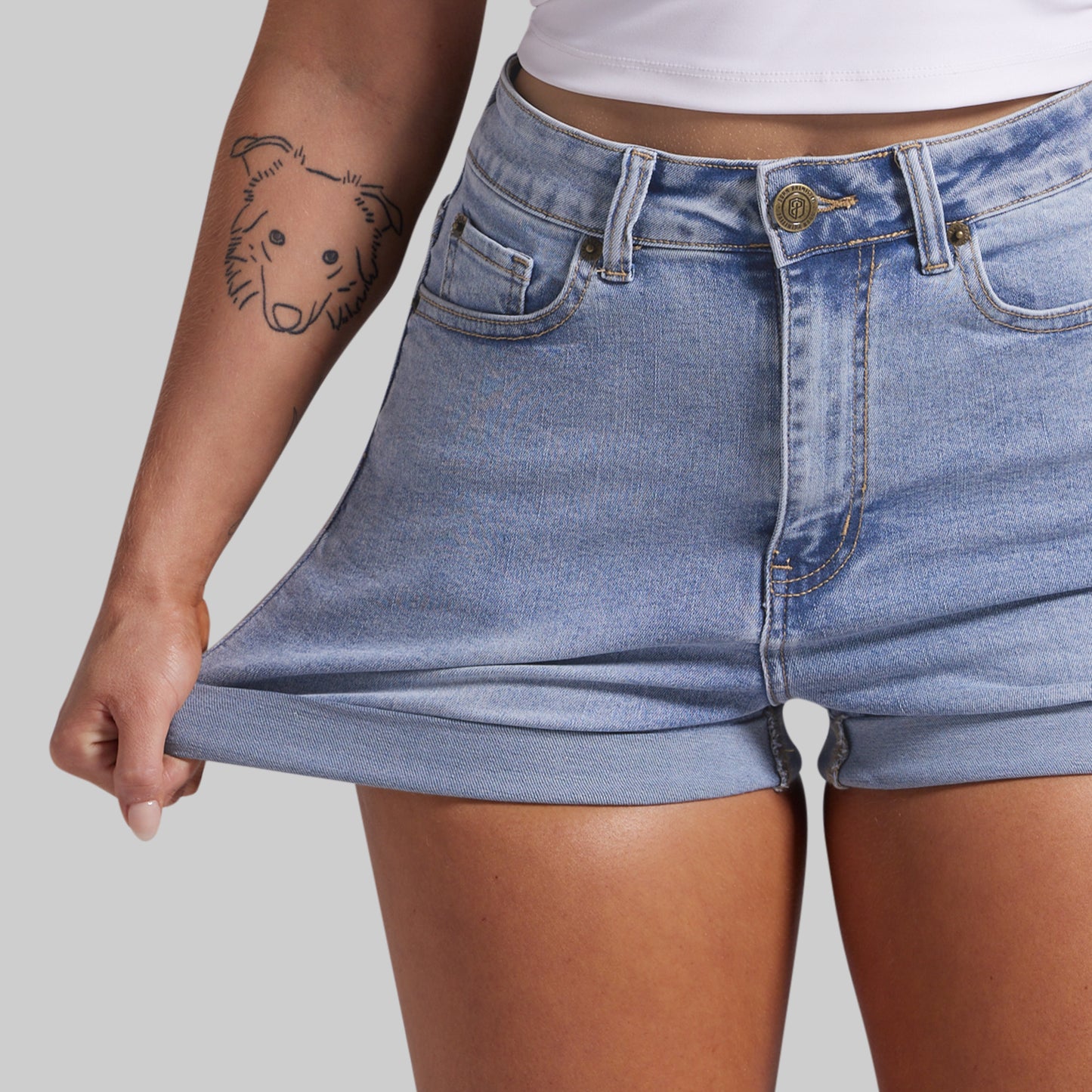Close-up of a person wearing light-wash blue denim shorts, pulling the hem outward to show stretch. The person is also wearing a white cropped top against a light gray studio background.