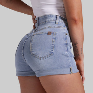 Close-up back view of a person wearing light-wash blue denim shorts with rolled hems and a small brown brand tab on the back pocket, against a plain light gray background.