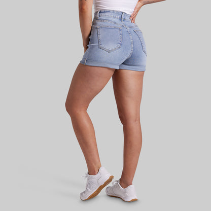 A model shown from the back wearing light-wash blue denim shorts, a white top, and white sneakers against a solid gray studio background.