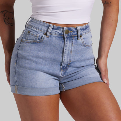 A close-up of a model wearing light-wash blue denim shorts with a rolled hem, paired with a white cropped tank top, against a solid gray background.