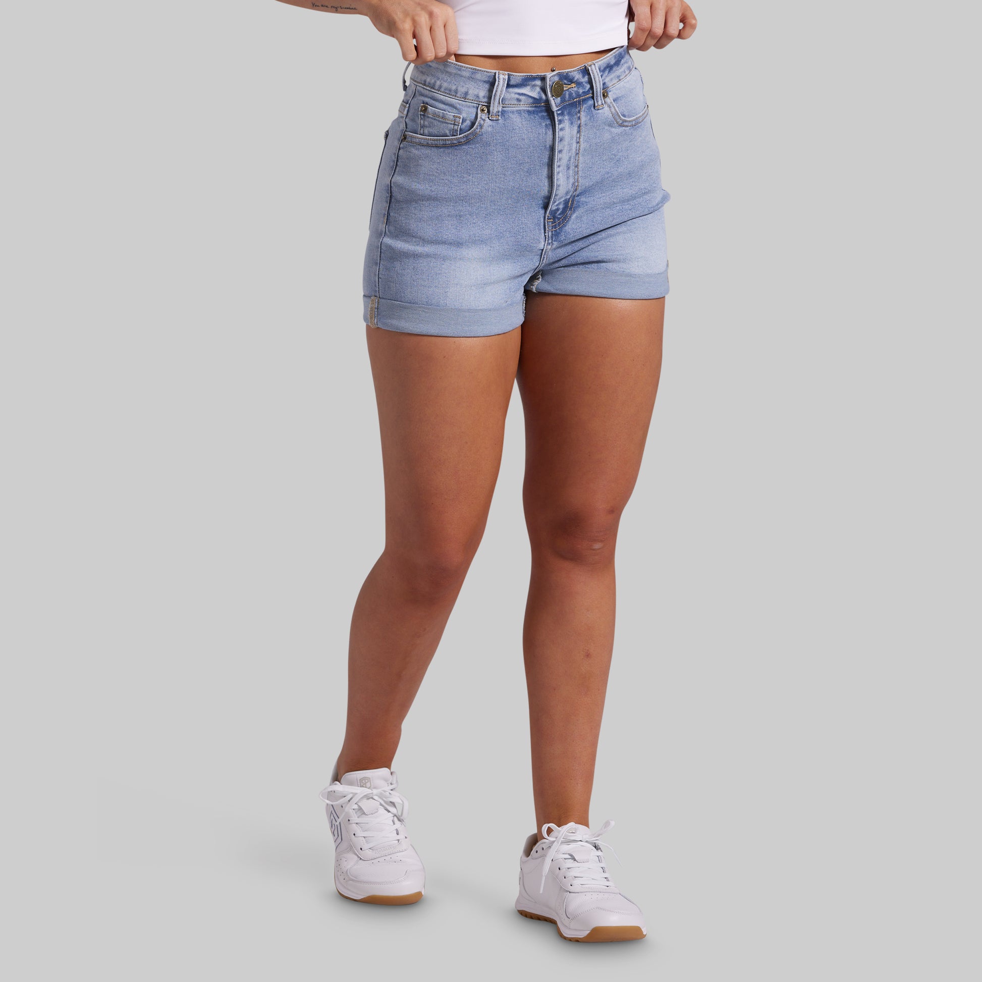 A model wearing light-wash blue denim shorts, a white crop top, and white sneakers, shown from the waist down against a solid gray background.