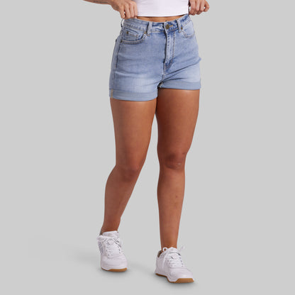 A model wearing light-wash blue denim shorts, a white crop top, and white sneakers, shown from the waist down against a solid gray background.