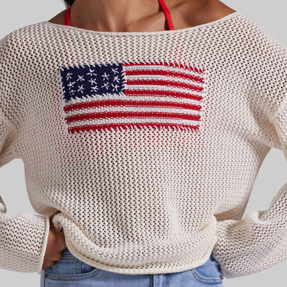 An off-the-shoulder knit sweater in cream with a large red, white, and blue American flag motif on the chest, worn by a model with blue jeans.