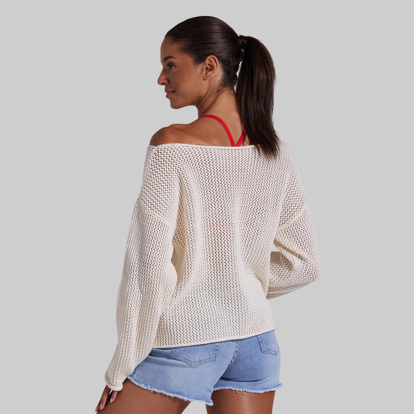 A woman shown from the back wearing an off-the-shoulder cream knit sweater and light blue denim shorts against a solid gray background.
