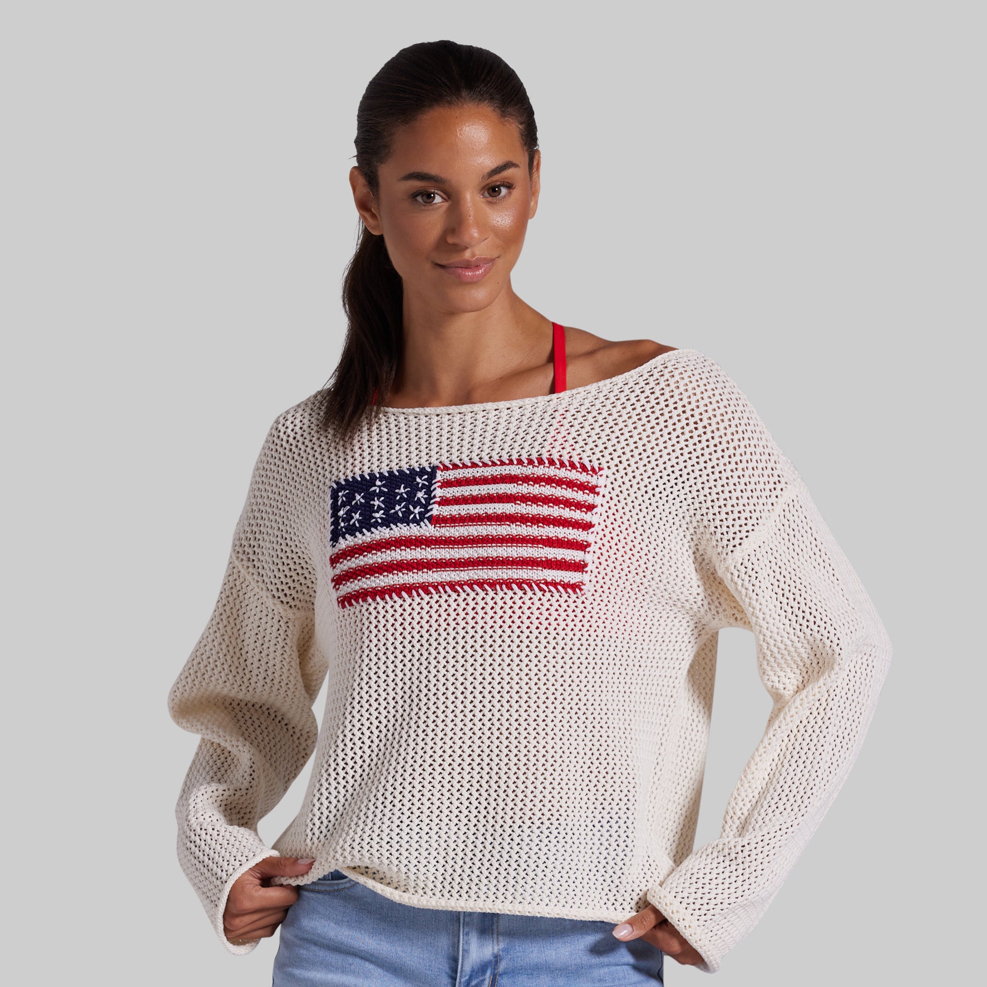 A woman wearing a cream-colored, open-knit long-sleeve sweater with a red, white, and blue American flag graphic on the chest, paired with blue jean shorts against a solid gray background.