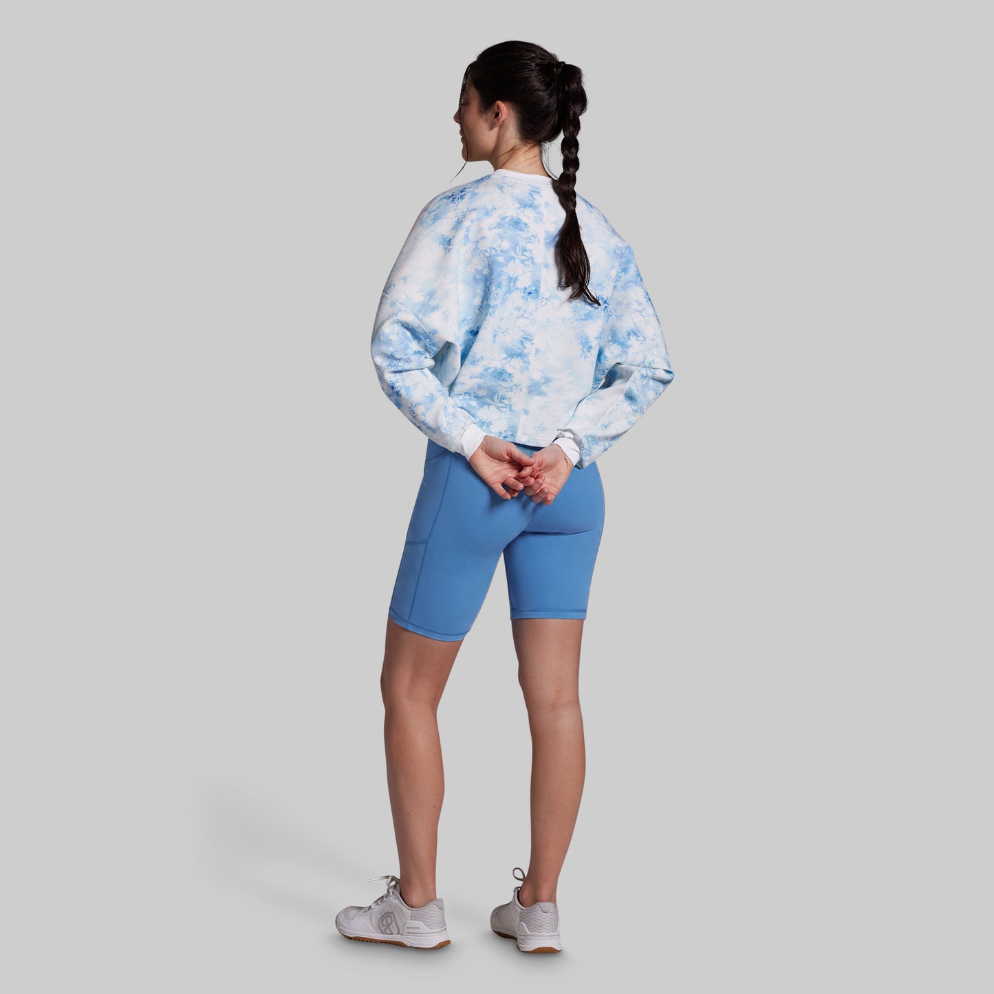A woman shown from the back wearing a light blue and white tie-dye long-sleeve top, blue fitted biker shorts, and white sneakers, standing against a solid gray background.